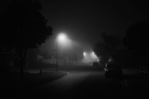 A black-and-white residential street disappearing into dense night fog beneath streetlights