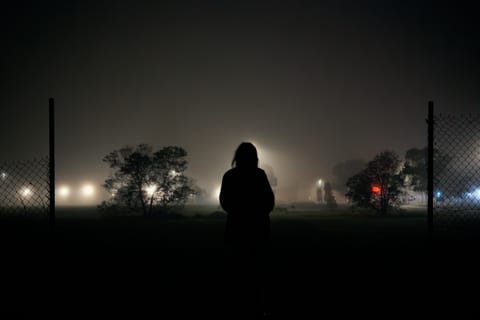 A silhouetted person standing between fences in a foggy, floodlit field at night